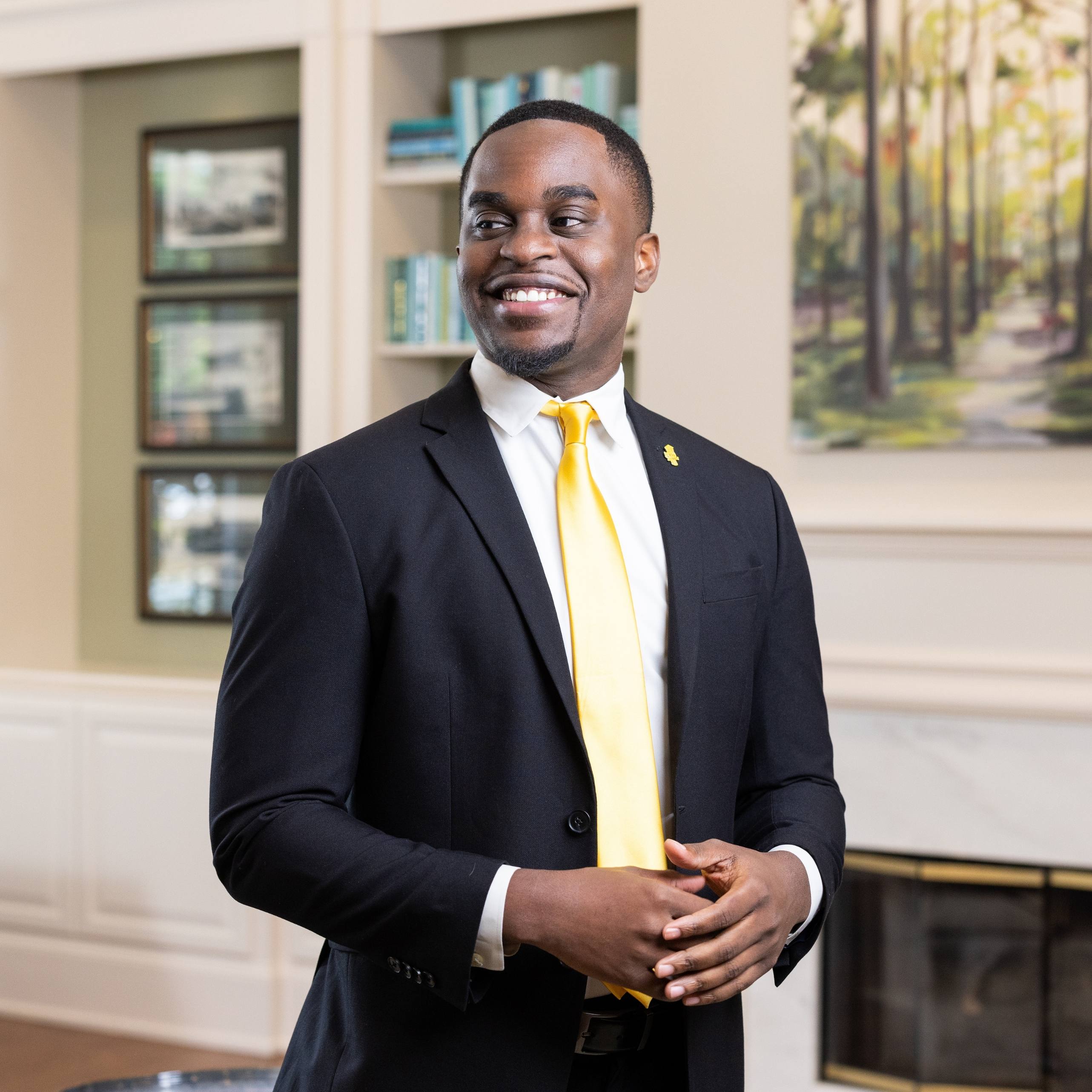 Person wearing a dark suit and gold tie standing indoors in front of bookshelves and a fireplace.