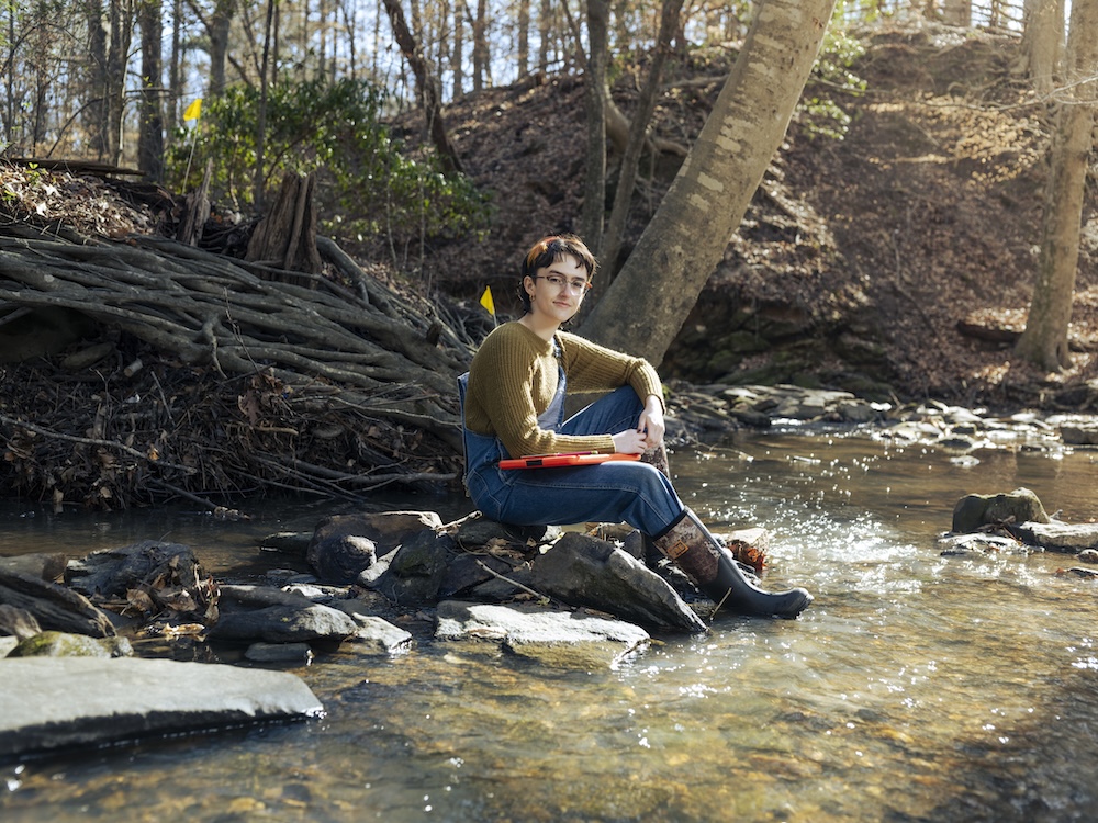 Student sitting in nature along a stream smiling for camera