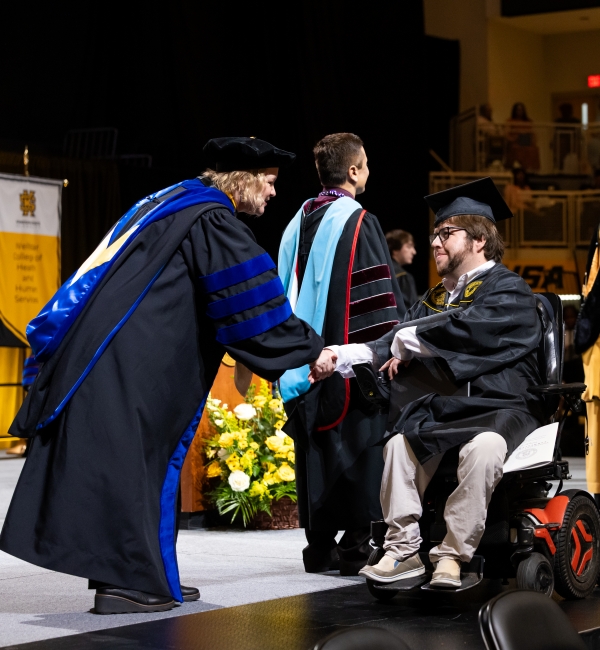 A graduate using an electric wheelchair receives a diploma from a faculty member in academic regalia during a commencement ceremony