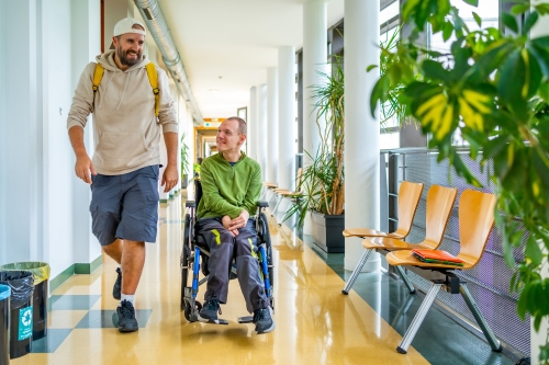 A person using a wheelchair converses with another person walking beside them in a hallway