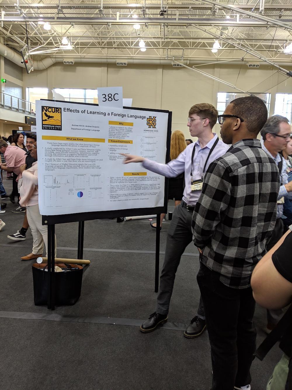 National Conference 2019 News / A young man presents a research poster titled "Effects of Learning a Foreign Language" to a small group at a bustling academic conference.