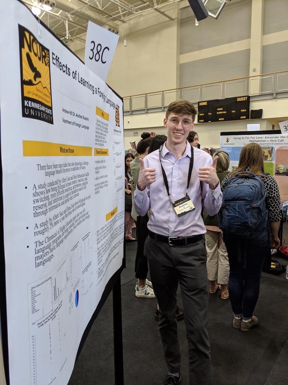 National Conference 2019 News / Young man in a light shirt gives a thumbs-up beside a university research poster on language learning. Background shows a busy academic conference.