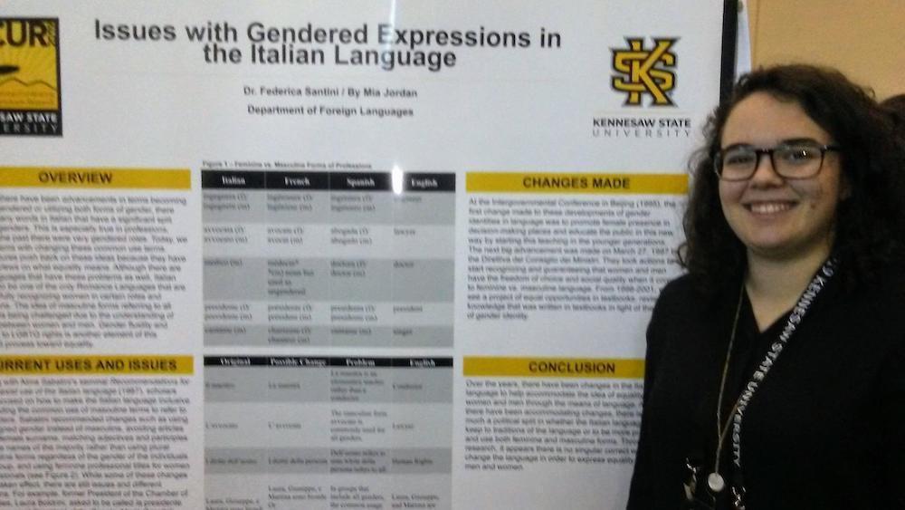 National Conference 2019 News / A person standing next to a research poster titled "Issues with Gendered Expressions in the Italian Language" at a Kennesaw State University event. The poster includes sections on overview, changes made, and conclusions. The individual is smiling, creating an informative and positive tone.