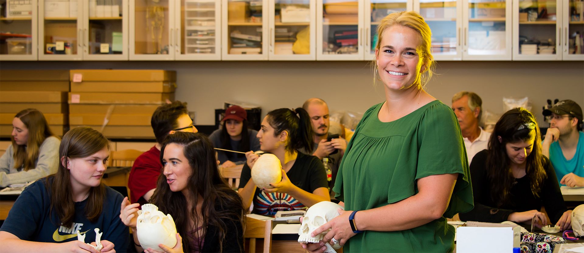 Smiling instructor holding a skull model in a classroom while students engage in hands-on learning.