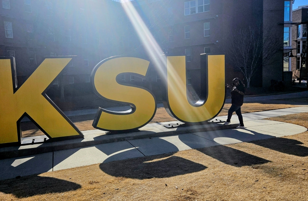 Large gold letters spelling “KSU” displayed outdoors on a sunny day, with a bright sunbeam shining diagonally across the scene. Alicia McGee standing to the right of the letters.