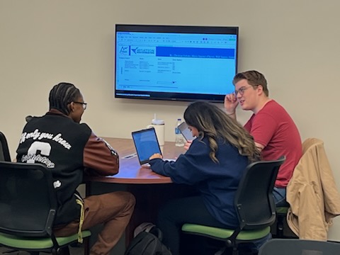 Three people seated around a round table in a meeting room. One person wears a black jacket with white text, another a navy blue hoodie working on a laptop, and the third a red shirt with glasses. A large screen behind them displays an Excel spreadsheet. The table holds water bottles and notebooks