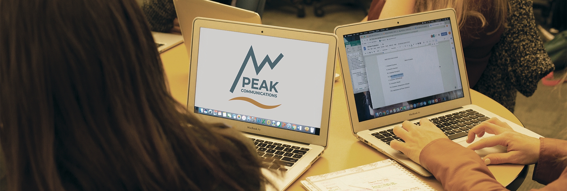 Two open laptops on a table. The left screen displays the Peak Communications logo with a stylized mountain and wave design, while the right screen shows a document being edited in Google Docs. Several people are seated around the table, collaborating.