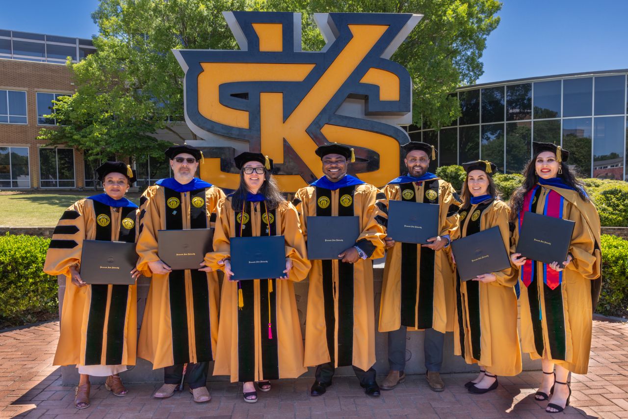 Spring 2026 Graduates of the CM Ph.dD program standing in cap and gown near KSU letters