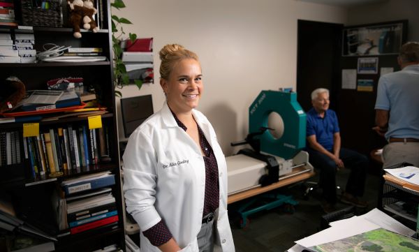 A smiling anthropologist in a lab coat stands in an office with bookshelves and equipment.