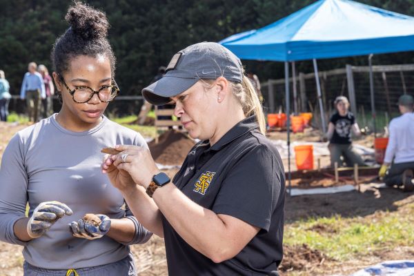 Two people at an archaeological dig site examining an artifact, with excavation work and team members in the background.
