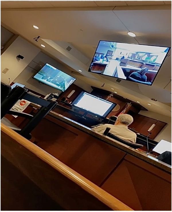 Inside a courtroom, people are seated at desks, focused on a television display in front of them
