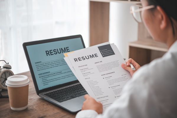 A person holding a resume in their hands, with a laptop sitting on a desk, displaying a resume.