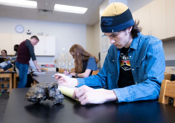 A student in a beanie and denim jacket takes notes while examining a rock specimen in a science classroom.