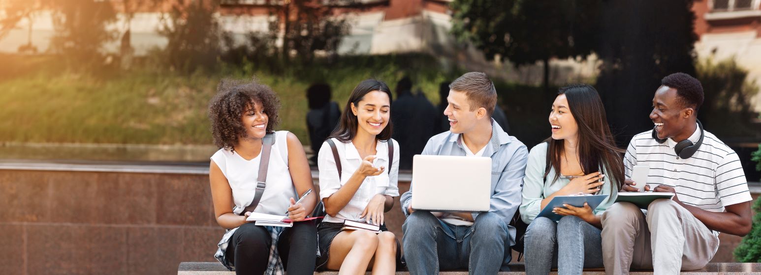 Cheerful college students resting outdoors between classes, using laptop, chatting and laughing.