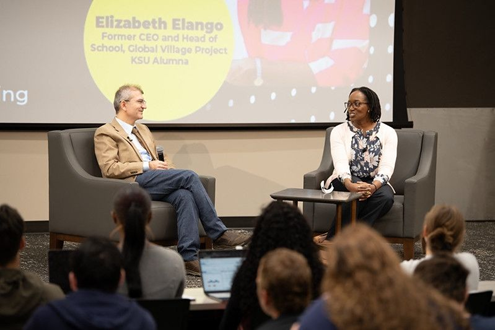 Ramazan Kilinc interviewing Elizabeth Elango both are sitting in large grey chair on a carpeted stage with a large serrn behind