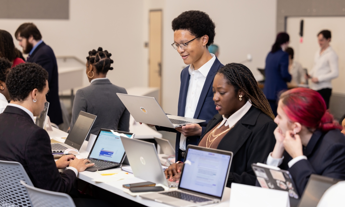 Kennesaw State students in suits sit at a long classroom table with laptops open. One student stands in the middle.