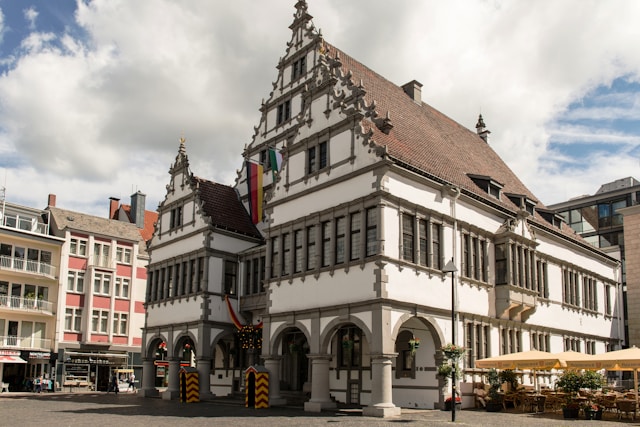 Historic white Renaissance-style town hall with ornate stepped gables, arched arcades, and red tiled roof beside pink buildings, flags flying under cloudy sky.