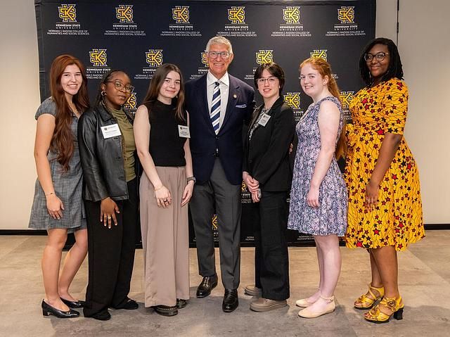 Group of seven people posing in front of a Kennesaw State University backdrop, including students and a man in a suit.