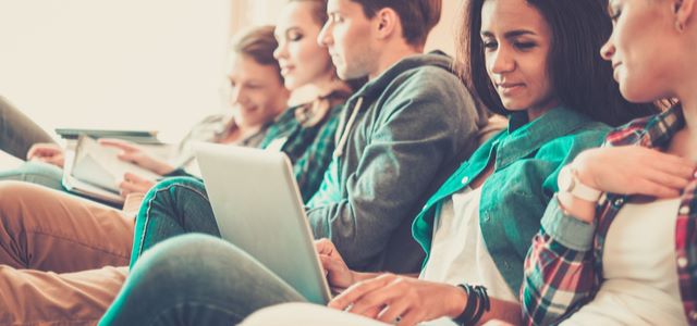 A diverse group of young individuals seated on a couch, each engaged with their laptops and notebooks.