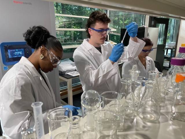 Three lab technicians wearing white coats, safety goggles, and blue gloves work with glassware and a pipette in a laboratory.
