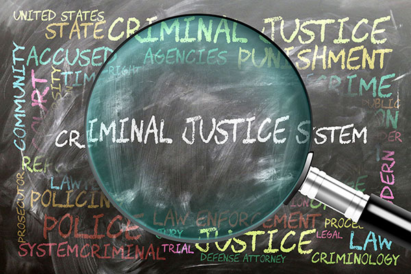 Magnifying glass highlighting the words "Criminal Justice System" on a chalkboard filled with related terms such as law enforcement, prosecution, trial, and punishment.