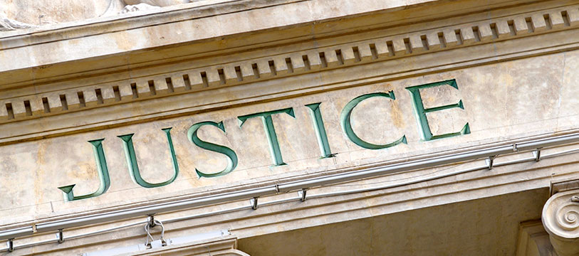 A close-up photograph of the word "JUSTICE" engraved in large, green capital letters on the facade of a classical-style stone building