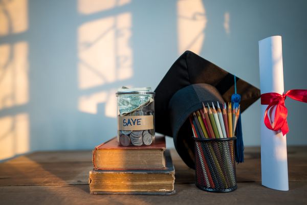 A graduation cap, diploma, pencils, two books, and a jar of money with the word "save" on it, displayed on a wooden table.