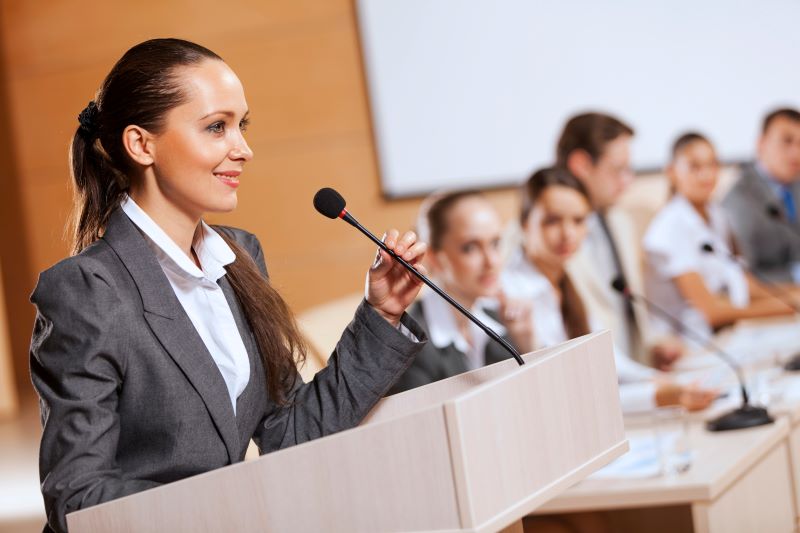 A woman holding a microphone, standing at a podium.