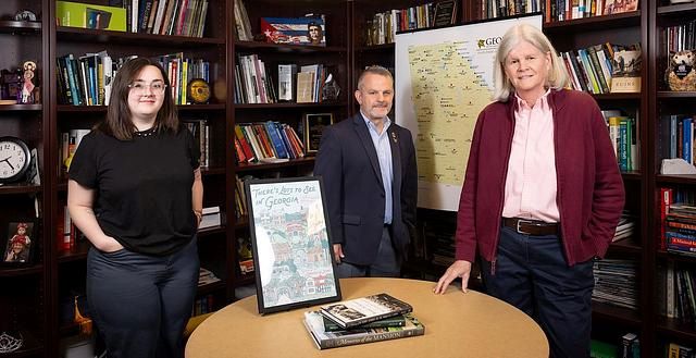 Three people standing in a library-like room with bookshelves, a round table with books, and a Georgia-themed poster and map in the background.