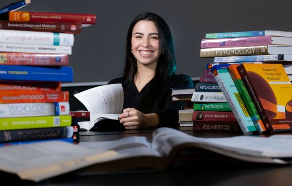 A woman smiling with a book in her hand, surrounded by piles of Psychology textbooks.