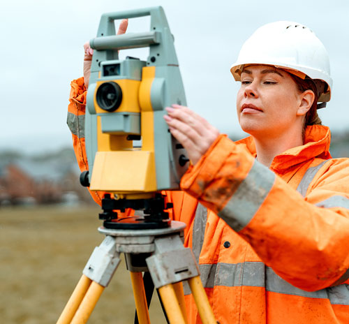 Geospatial Sciences professional wearing a hard hat and high-visibility jacket, operating a surveying total station instrument on a tripod outdoors.