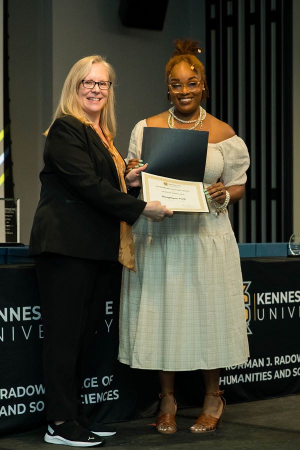 Two women standing in front of a stage, holding a certificate