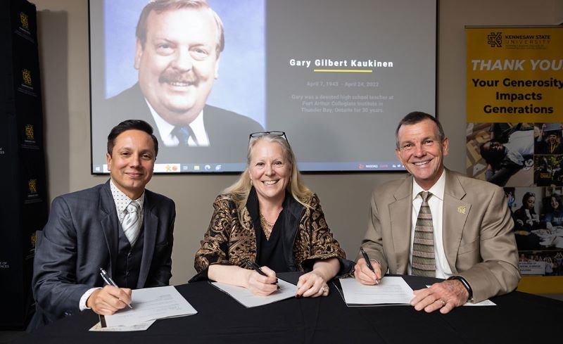 Ivan Pulinkala, Katie Kaukinen, and Lance Burchett, sitting at a table with pens in their hands, signing a document.