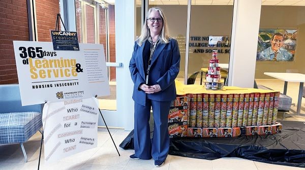 A woman in a blue suit stands next to a display about housing insecurity, with canned goods arranged on a table behind her.