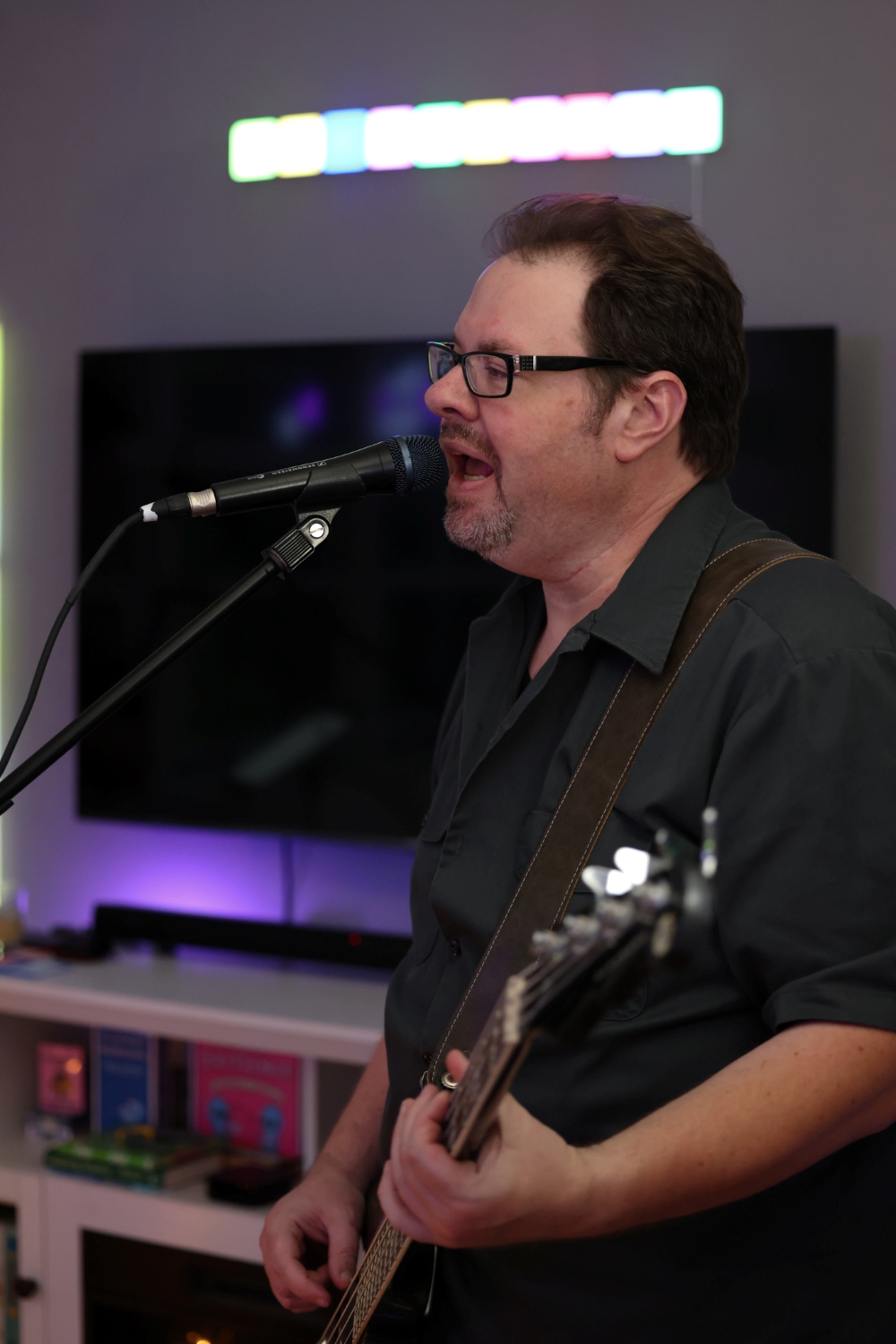 Tim Kersey with glasses and short hair singing into a microphone while playing an electric bass guitar. The background includes a television screen and colorful lighting