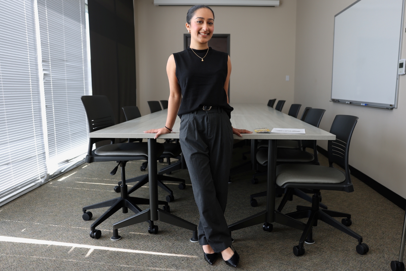 Diya Patel wearing a coral-colored dress with layered pearl necklaces and a patterned shawl, standing with arms crossed in front of a brick wall.