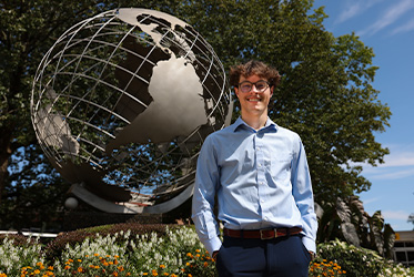 Christian Sousa wearing a light blue dress shirt and dark trousers stands outdoors in front of a large metallic globe
