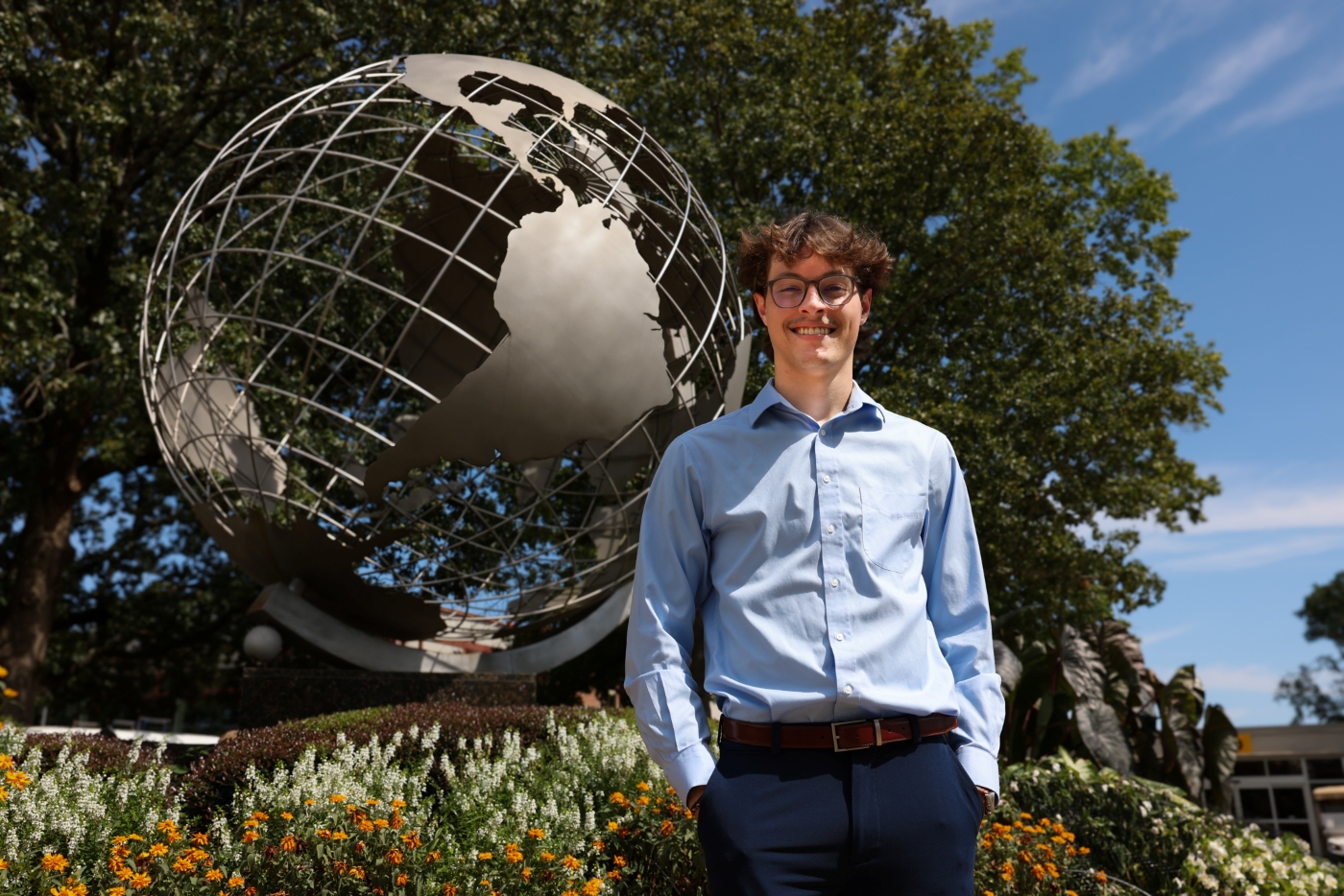 Christian Sousa wearing a light blue dress shirt and dark trousers stands outdoors in front of a large metallic globe
