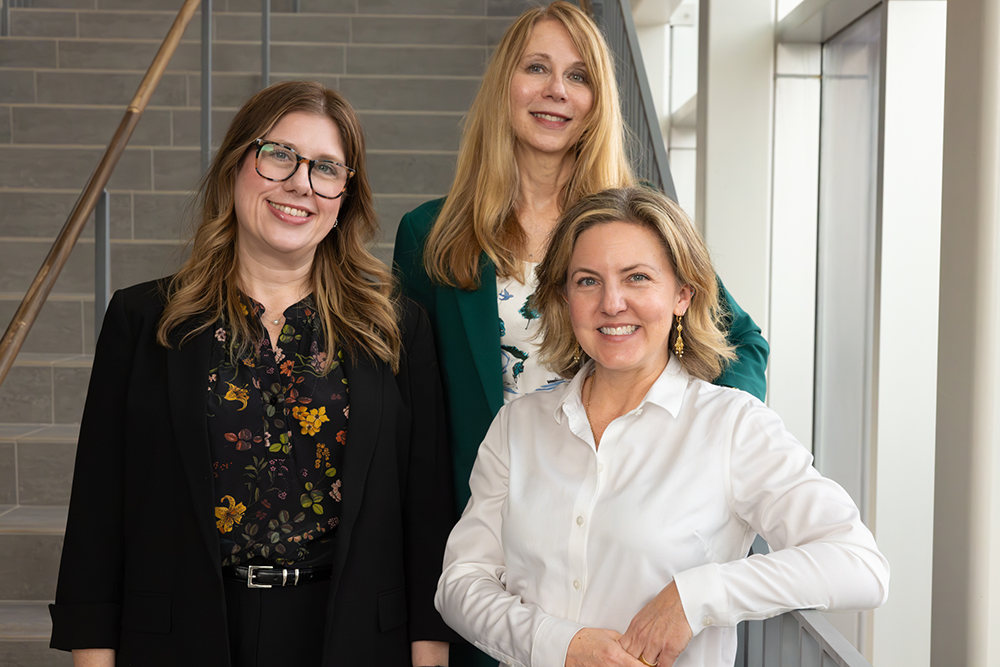 Three professionals stand together on an indoor staircase, dressed in business‑casual attire, with natural light from nearby windows in a modern building.