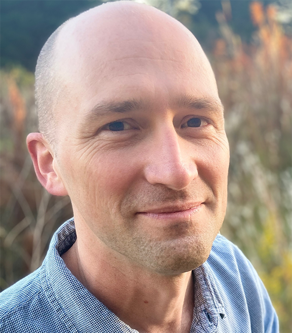 Dr Tom Okie headshot outdoors, wearing a blue button-up shirt, smiling gently, with a blurred natural background of greenery and tall grasses.