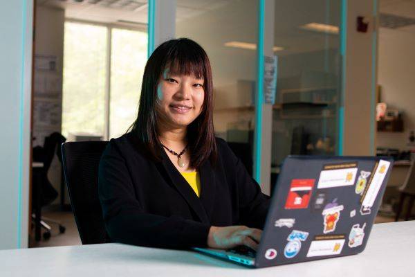 A woman smiling while using a laptop, in a classroom setting.