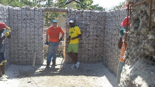  / Two individuals stand in a partially constructed building with walls made of wire mesh filled with rocks.
