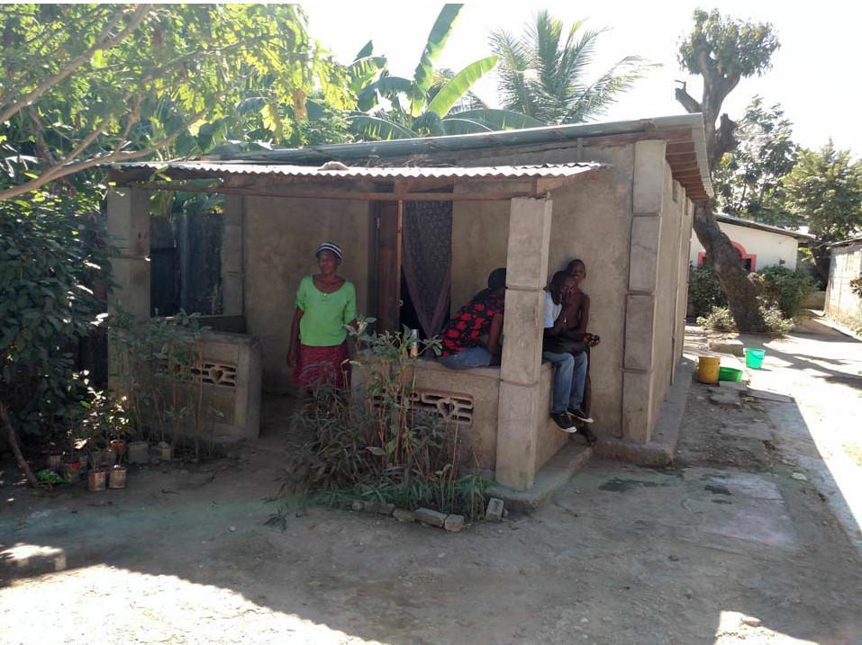  / A small concrete house with a tin roof and open porch, surrounded by greenery