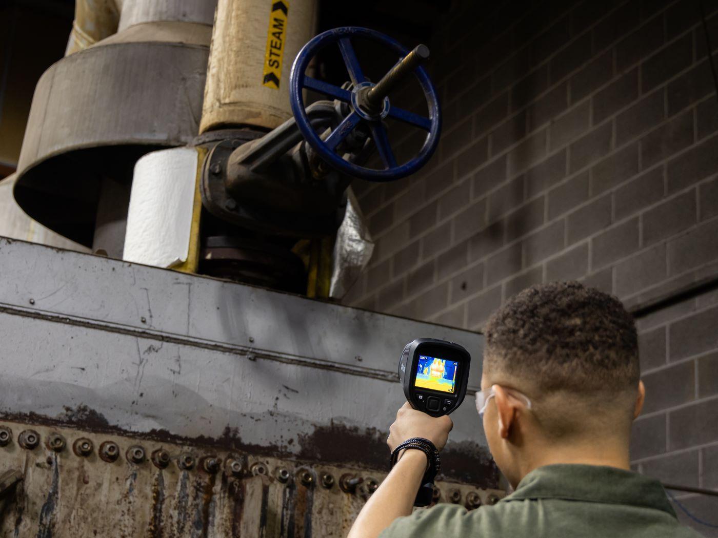  / Person using a thermal imaging camera to inspect a pipe with a blue valve labeled "STEAM." 
