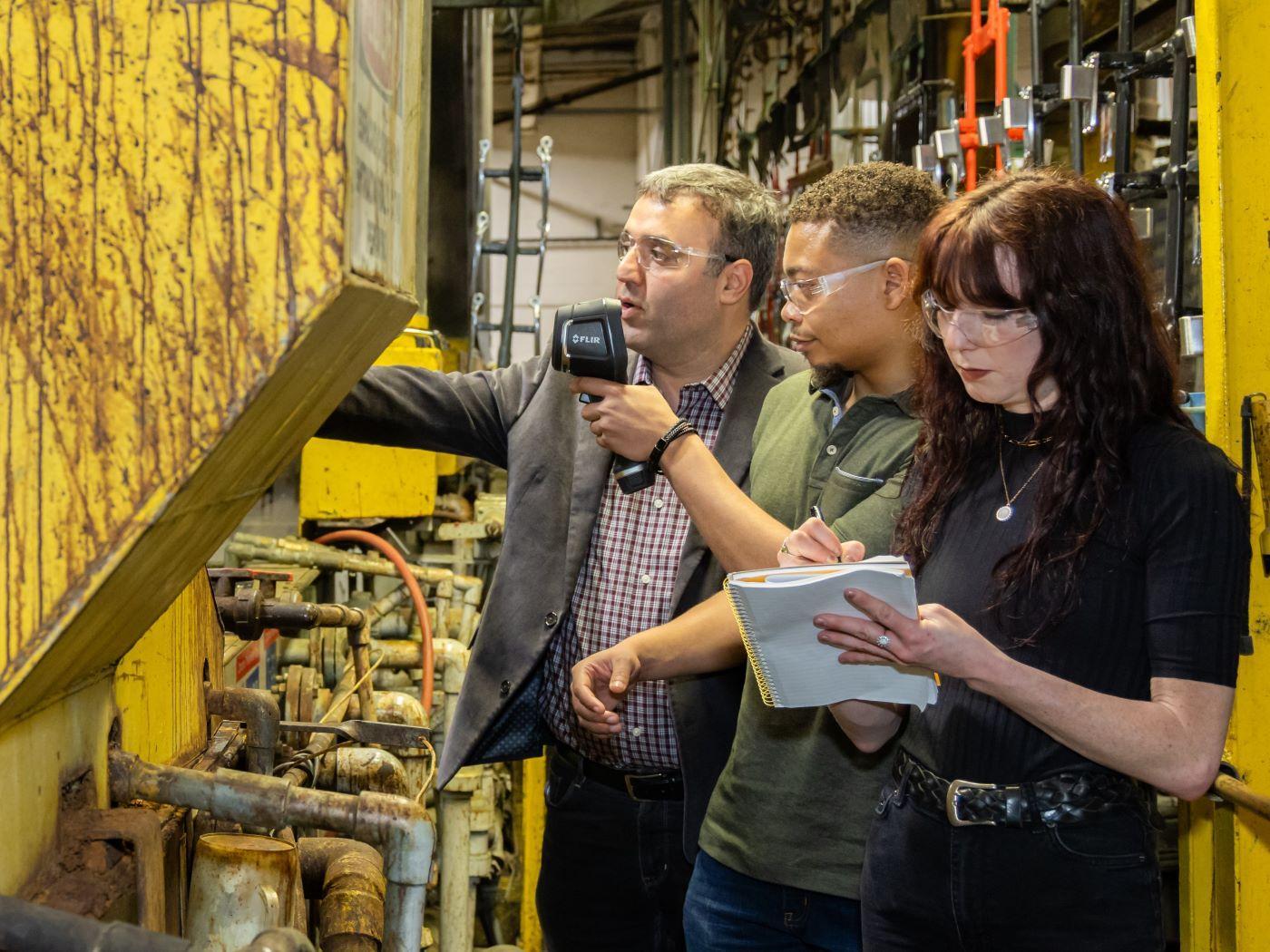  / Three people inspecting yellow industrial machinery, one holding a device and another taking notes. 
