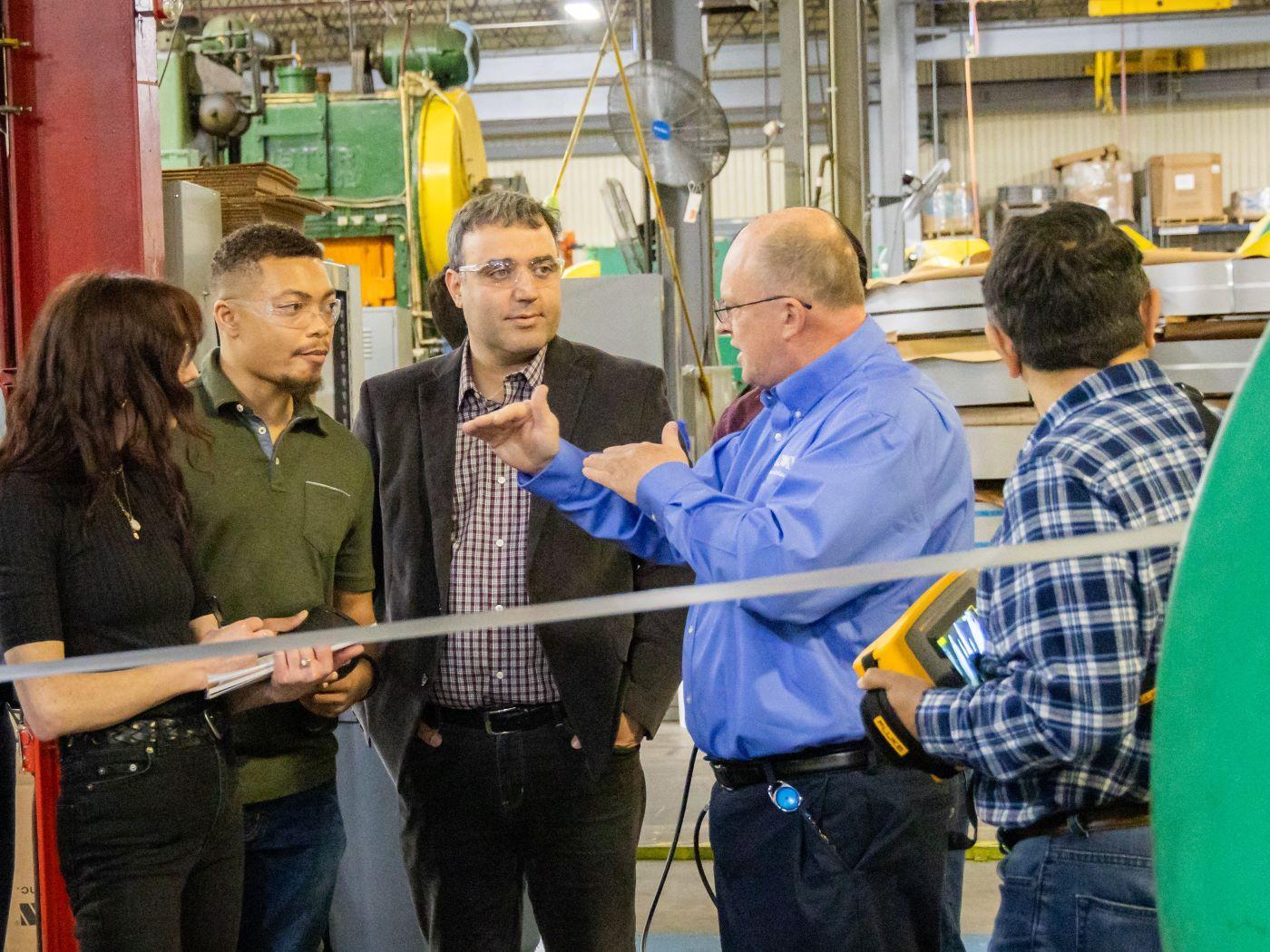  / Five people standing in an industrial setting, engaged in discussion. 

