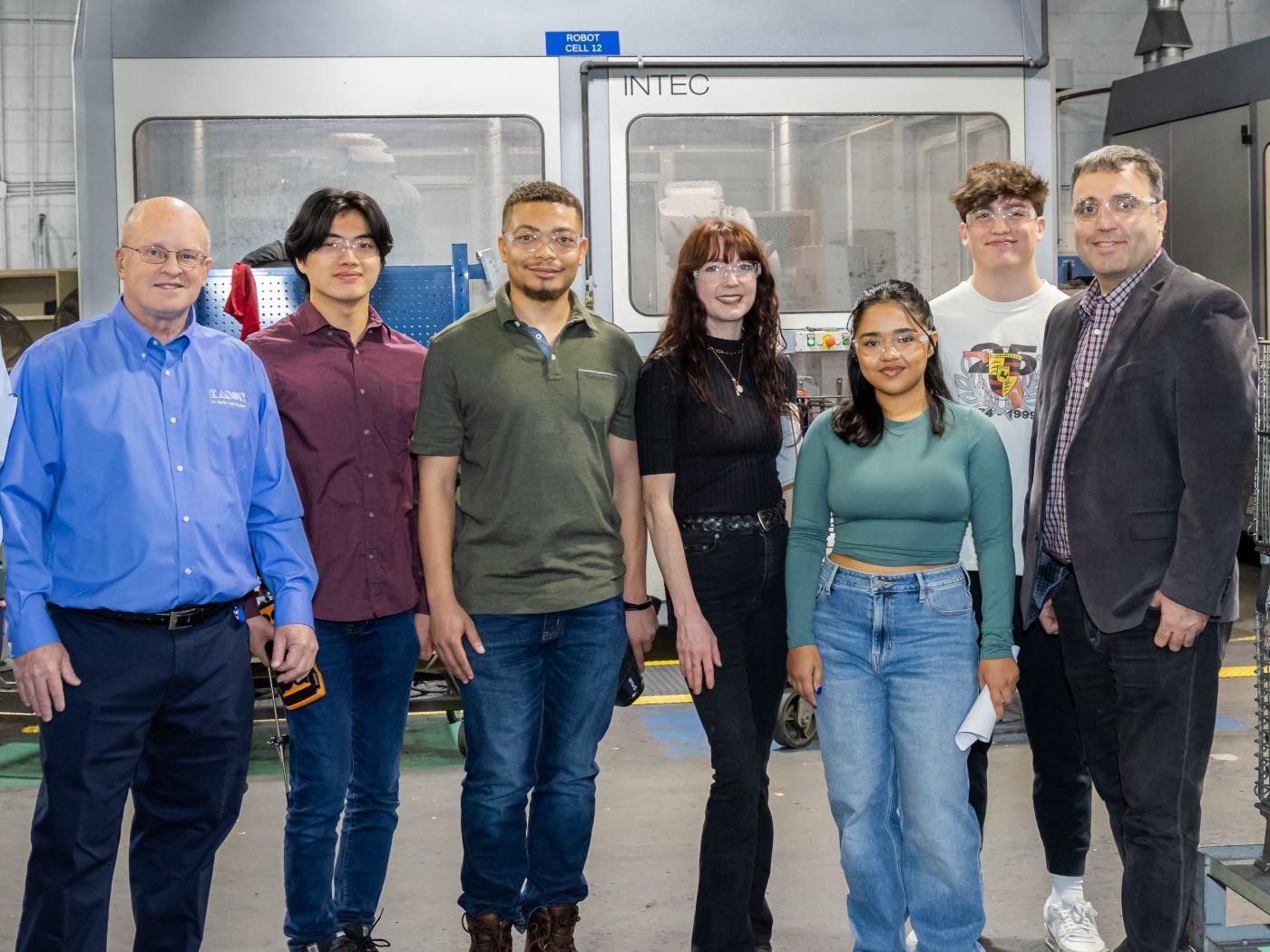  / Seven people standing in front of industrial machinery in a factory. 

