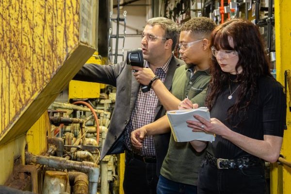 Three people in a factory setting, with one person holding a device and another taking notes.