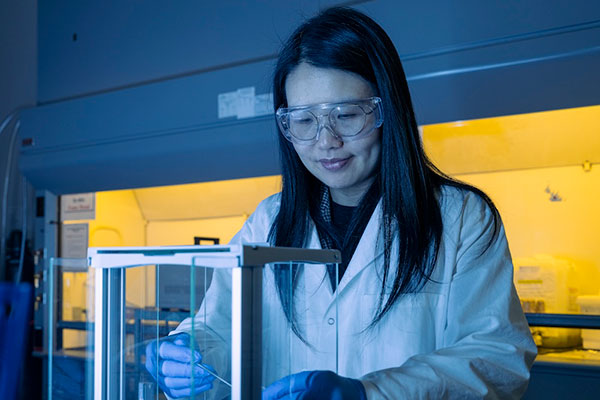 Beibei Jiang in a lab coat and safety goggles works in a science lab with a focused expression. She handles a sample inside a yellow-lit fume hood.