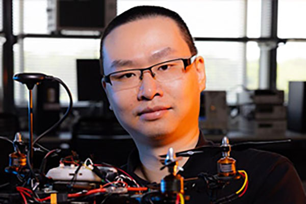 Yan Fang wearing glasses is holding a drone, standing in a tech-filled lab. The atmosphere is focused and innovative, highlighting technology.